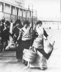 Black and white photograph of immigrants on a dock. There is a woman in the foreground of the photo holding several bags. There are four other people behind her, and a boat in the background.