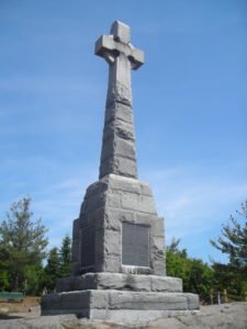 A tall memorial made out of gray stone in the shape of a cross, with a dark gray plaque at the base.
