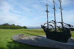 A photograph of the National Famine Monument, a statue of a coffin ship with three masts, resembling crosses, surrounded by skeletal figures